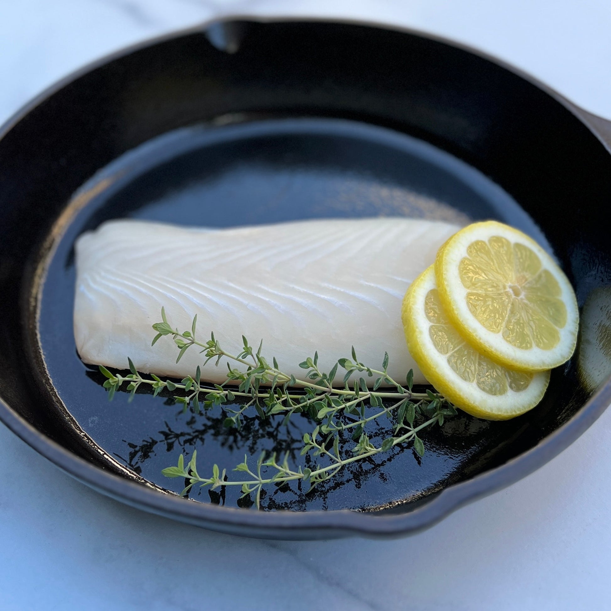 Raw halibut fillet in a cast iron pan with lemon slices and fresh thyme on a white background.