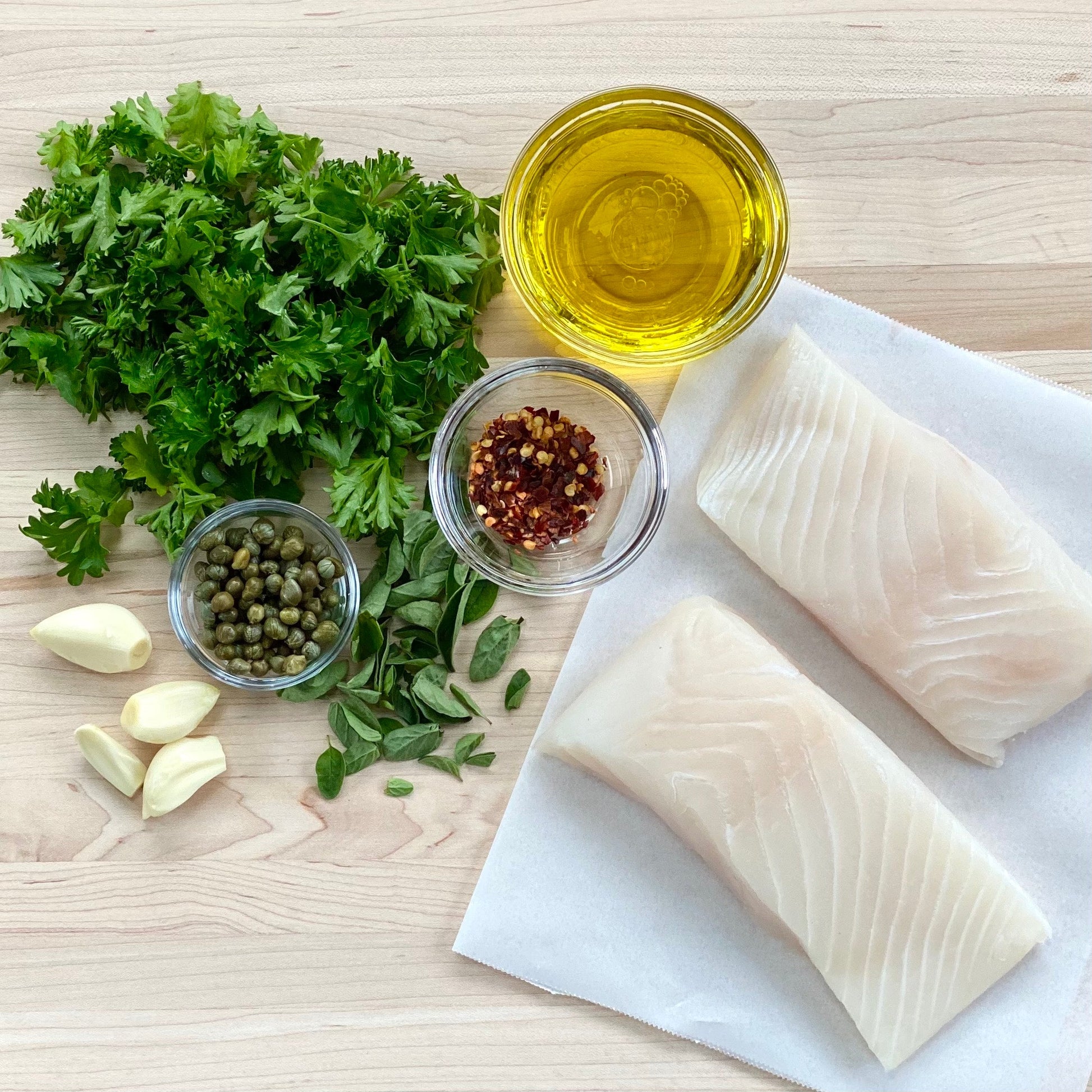 Two halibut portions on parchment with fresh herbs, garlic a small bowl of olive oil and red pepper flakes on top of a wood cutting board.