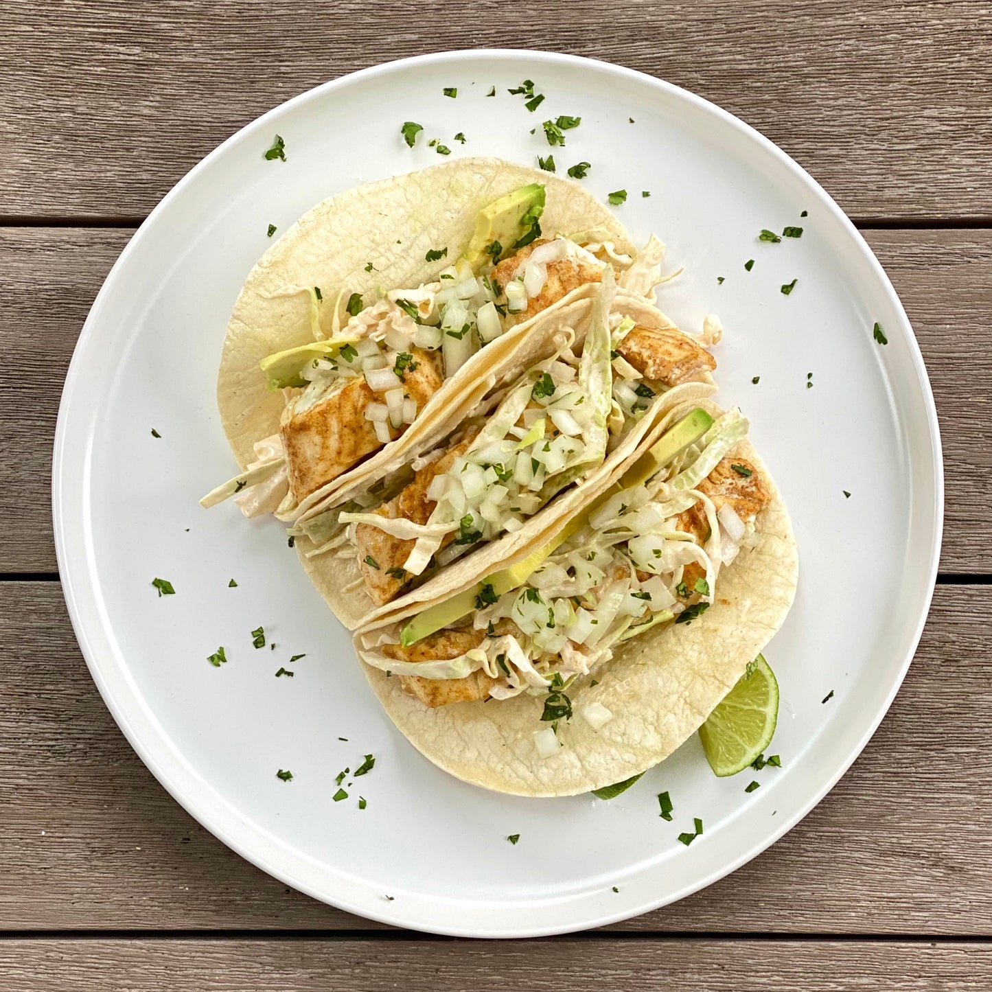 A plate with three halibut tacos with cabbage and a lime slice on top of a wood table.