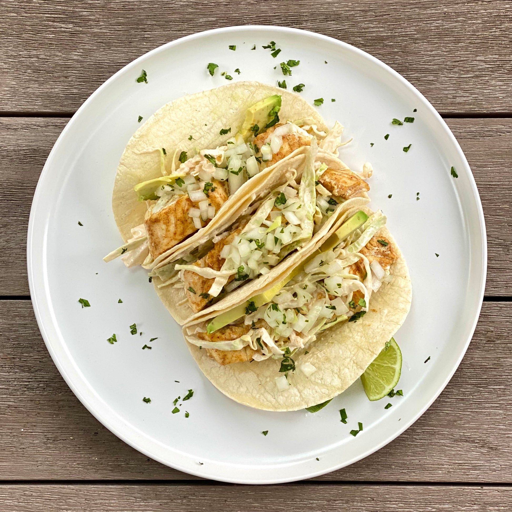 A plate with three halibut tacos with cabbage and a lime slice on top of a wood table.