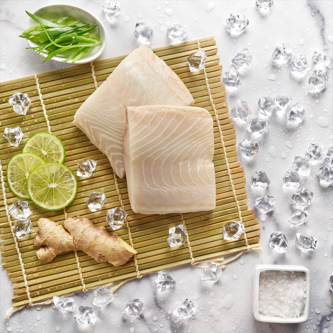 Wild sablefish fillets on a sushi mat with fresh ginger, lime slices, a bowl of salt and ice on a light marble background