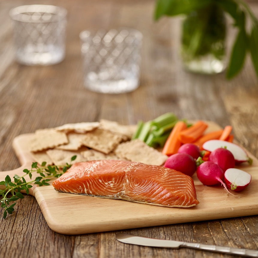 Wild Alaskan smoked king salmon fillet served on a wooden board with crackers, fresh radishes, carrots, and herbs.