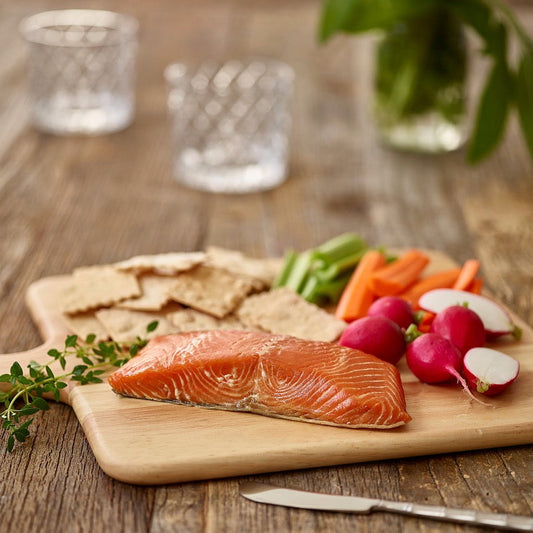 Wild Alaskan smoked king salmon fillet served on a wooden board with crackers, fresh radishes, carrots, and herbs.