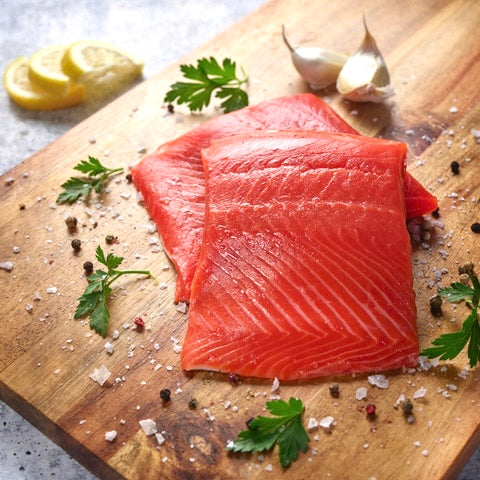 Two portions of sockeye salmon on a cutting board with fresh herbs, salt and pepper, garlic cloves and lemon slices in the background.