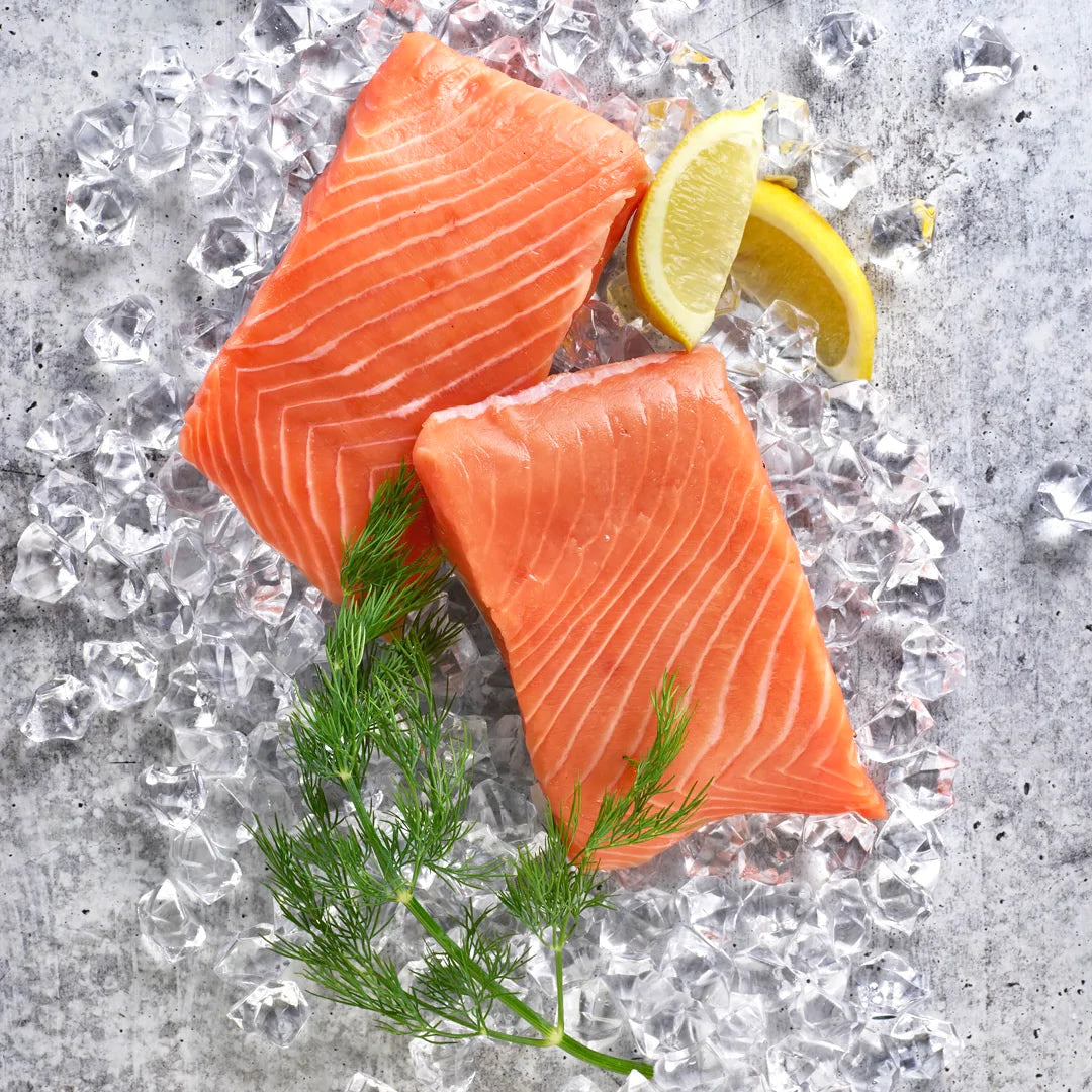 Two portions of wild alaskan king salmon on top of ice with two lemon wedges and a sprig of dill.
