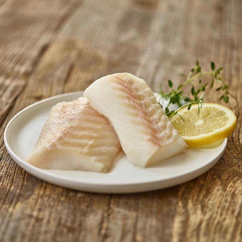 Two pieces of raw fish on a white plate with a lemon wedge and thyme sprigs on a wooden surface.