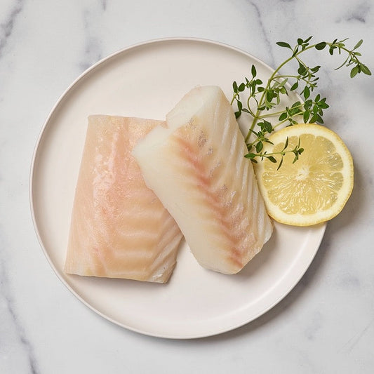 Two portions of wild alaska cod on a white plate with a slice of lemon and a sprig of thyme, presented on a marble surface.