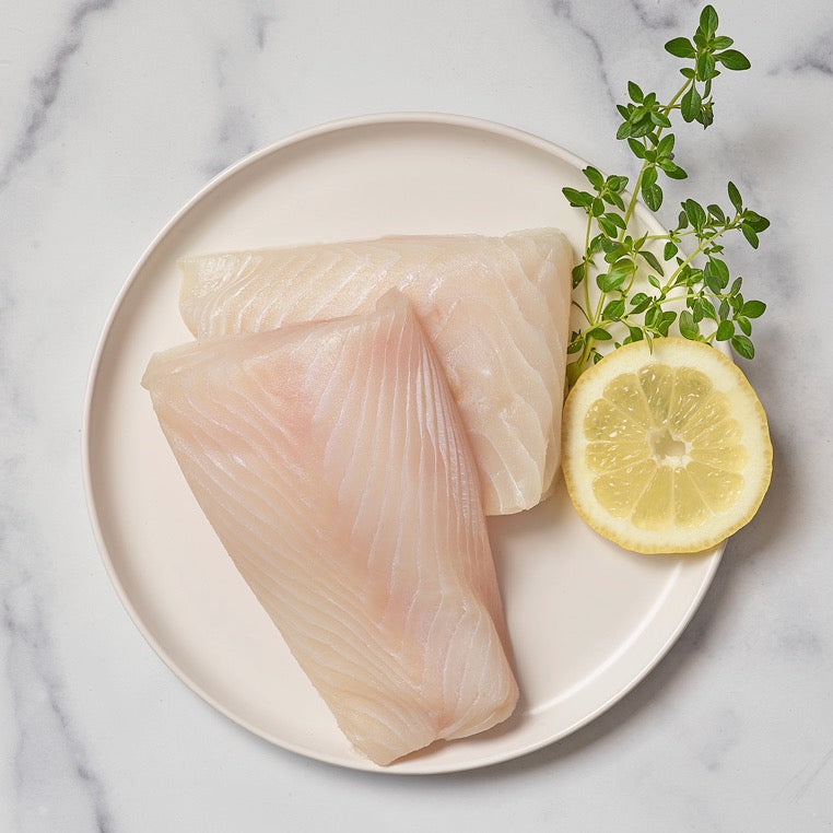 Raw halibut fillets on a plate with lemon and herbs on a marble background