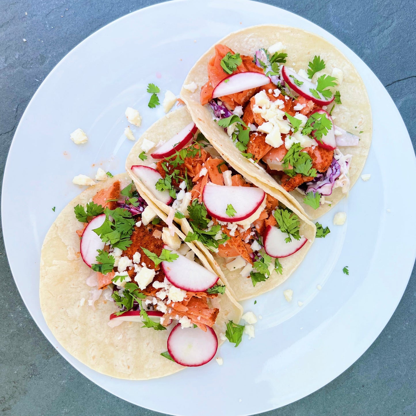 Salmon taco with radishes and fresh herbs and cheese on a white plate over a grey background.
