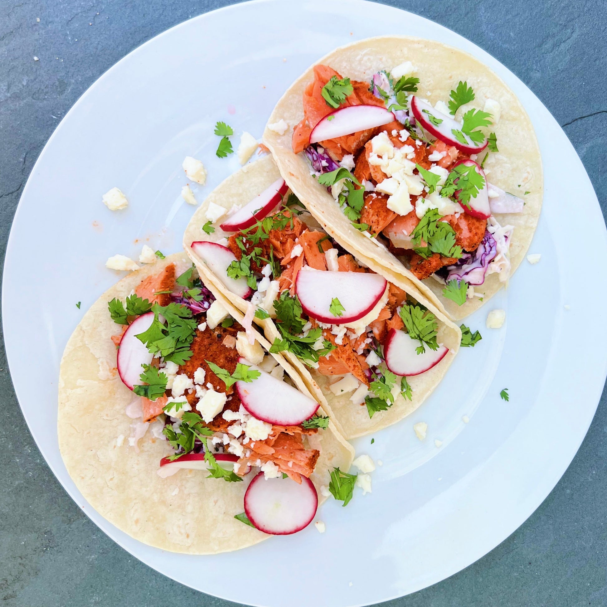 Salmon taco with radishes and fresh herbs and cheese on a white plate over a grey background.